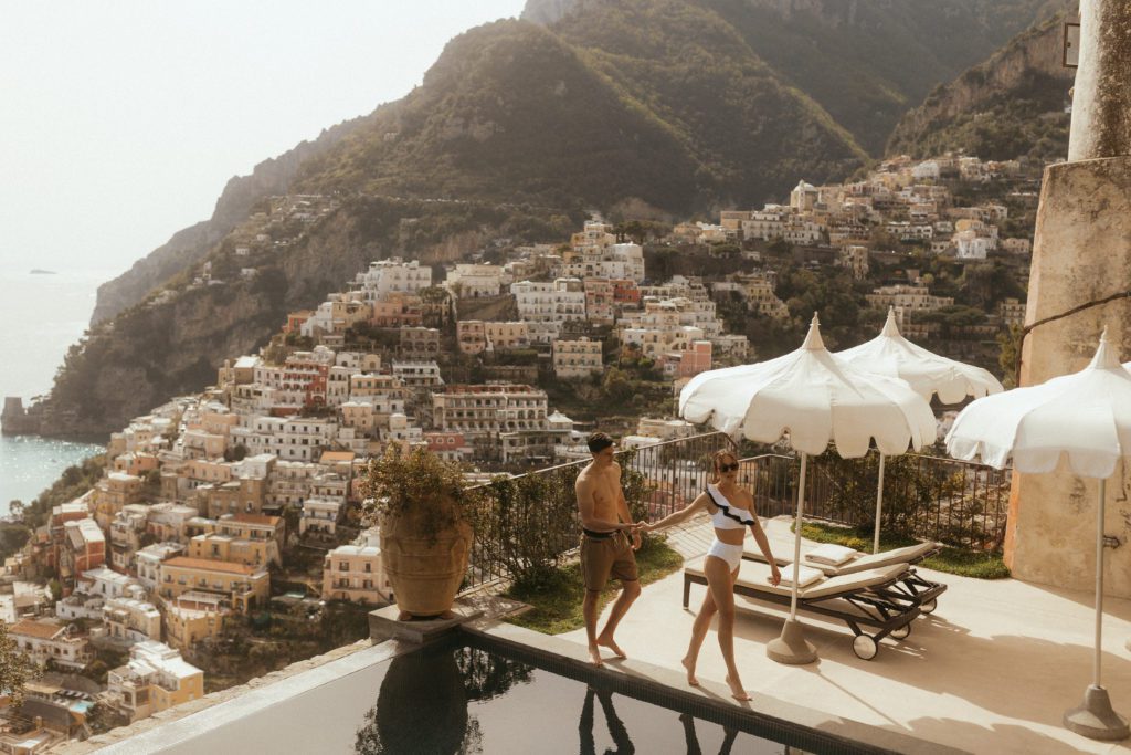 stress free travel tips, a couple in swim suits walk along the edge of a pool with a cliffside full of italian villas in the background